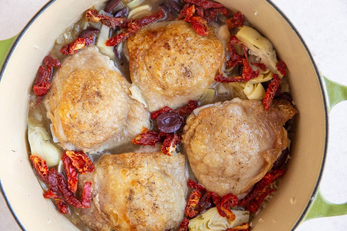 browned chicken thighs in a pot with sun-dried tomatoes, olives, artichoke hearts, rice, and more.