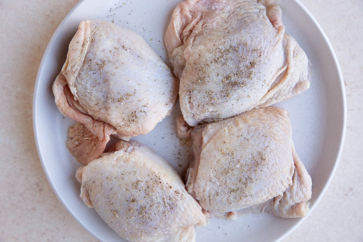 Raw chicken thighs seasoned with salt and pepper on a white plate.