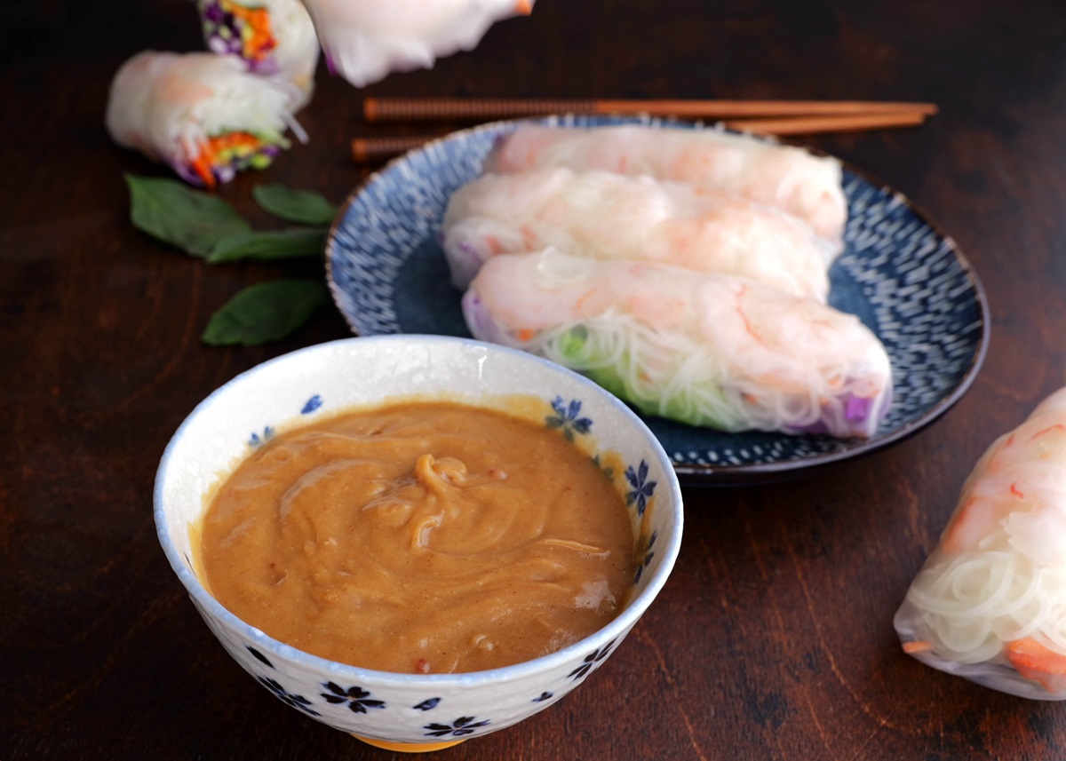 Bowl of peanut sauce with spring rolls in the background.
