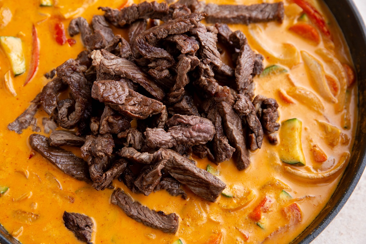 Steak added on top of red curry sauce with vegetables, ready to be mixed in.