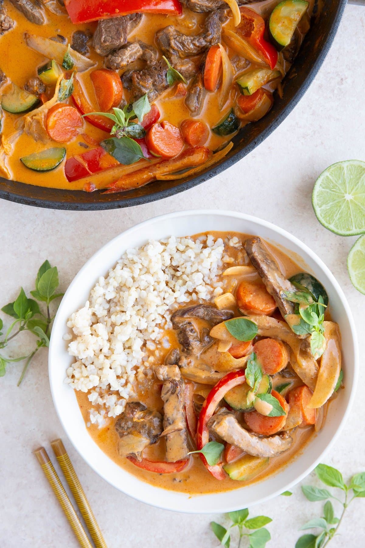 Bowl of Thai beef curry and skillet of Thai curry with fresh Thai basil and limes to the side.