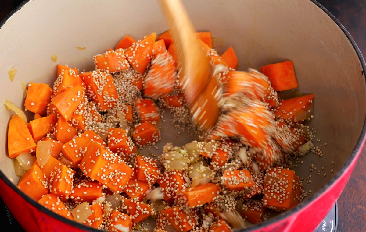 Quinoa being stirred into the pot with the onion and chopped sweet potatoes.