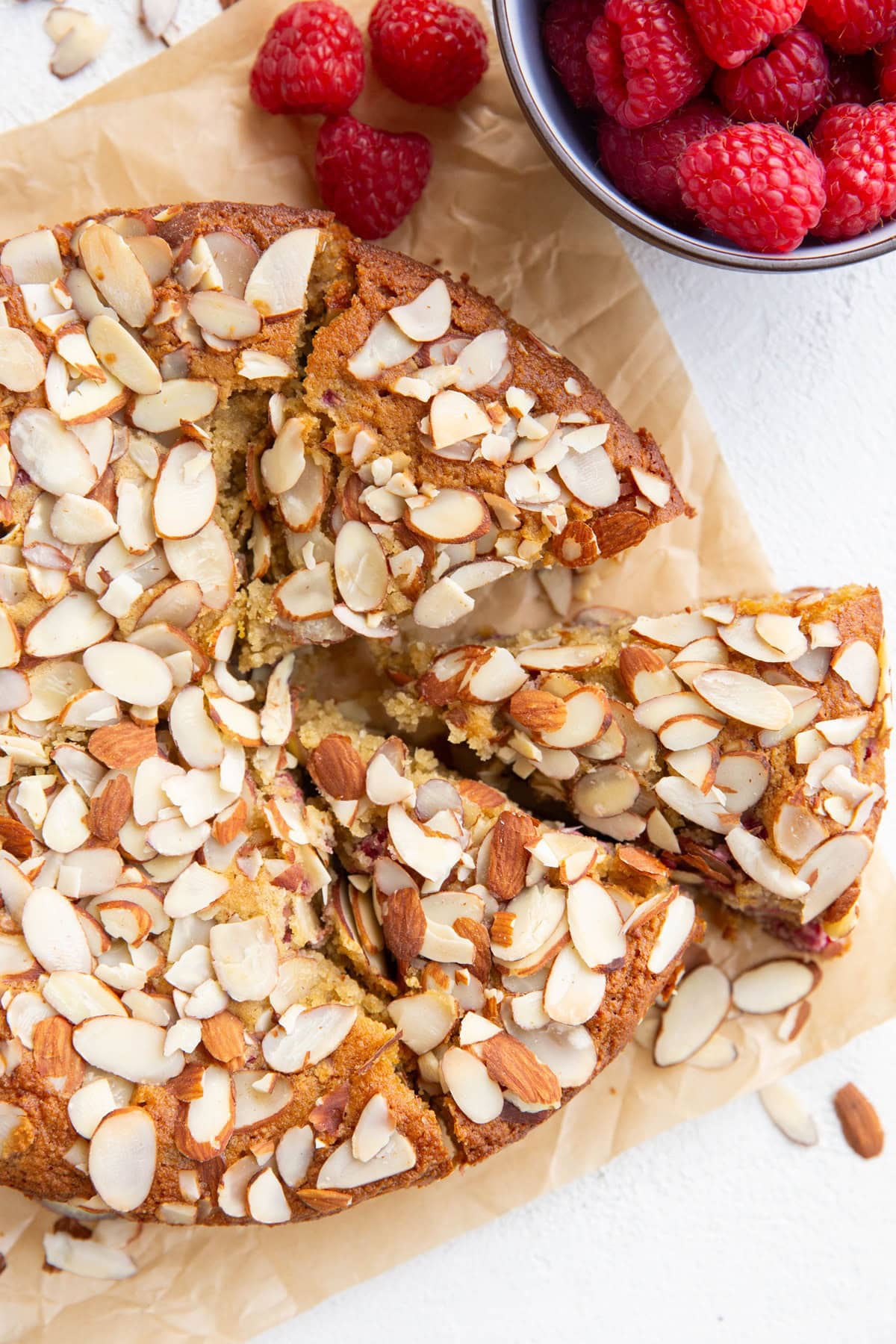 Full almond cake cut into slices with a bowl of fresh raspberries to the side.