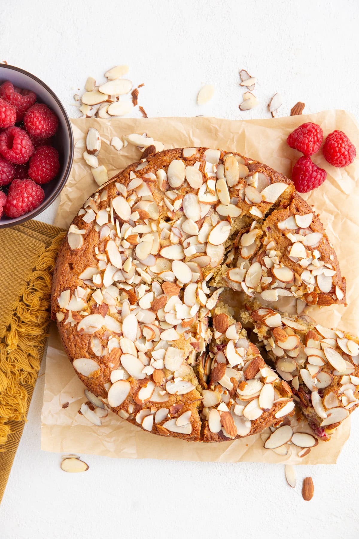 Raspberry almond cake on a white background with three slices cut. Fresh raspberries and sliced almonds all around.