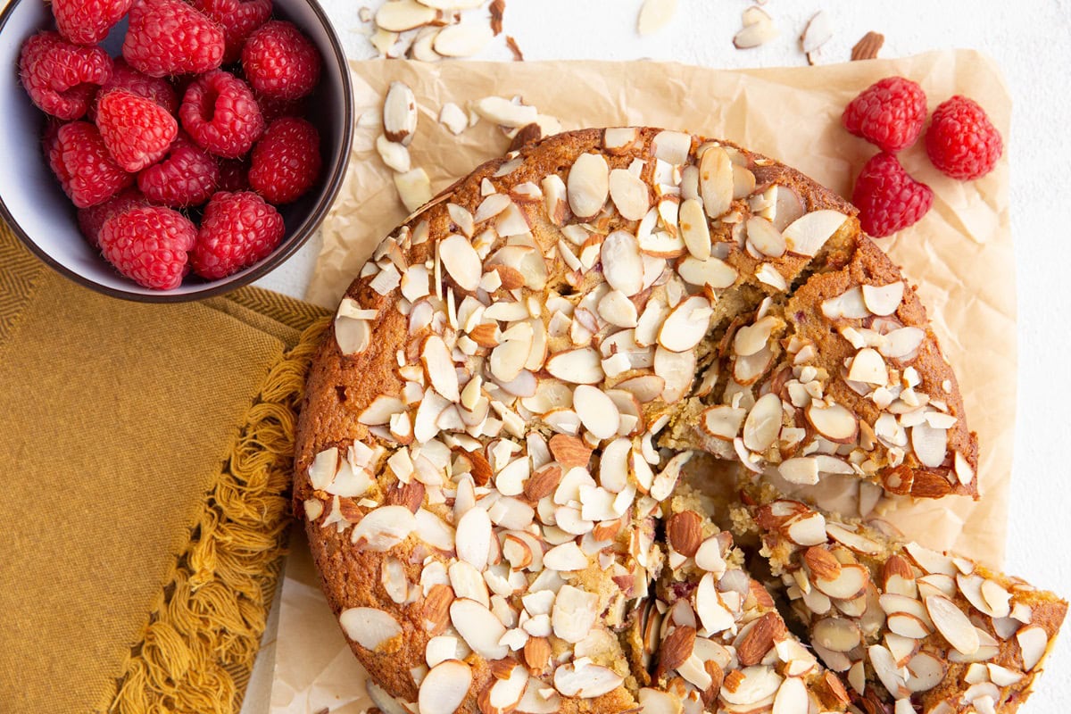 Whole cake cut into slices on a sheet of parchment paper with fresh raspberries around.