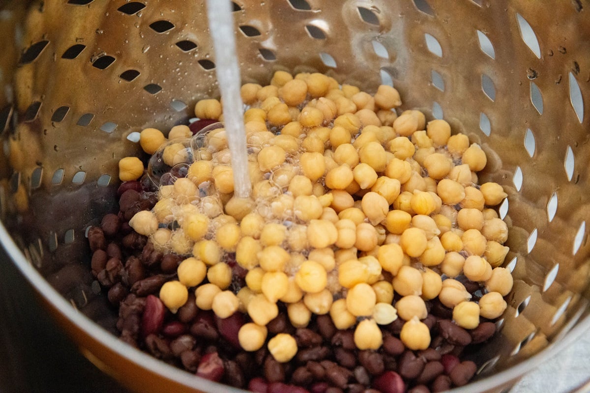 Colander with three types of beans inside, being rinsed with faucet water.
