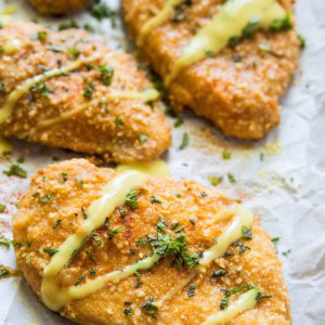 Four filets of Gluten-Free Baked Breaded Chicken on a large baking sheet, drizzled with sauce and sprinkled with fresh parsley.