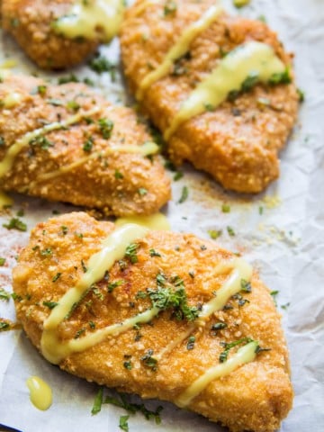 Four filets of Gluten-Free Baked Breaded Chicken on a large baking sheet, drizzled with sauce and sprinkled with fresh parsley.