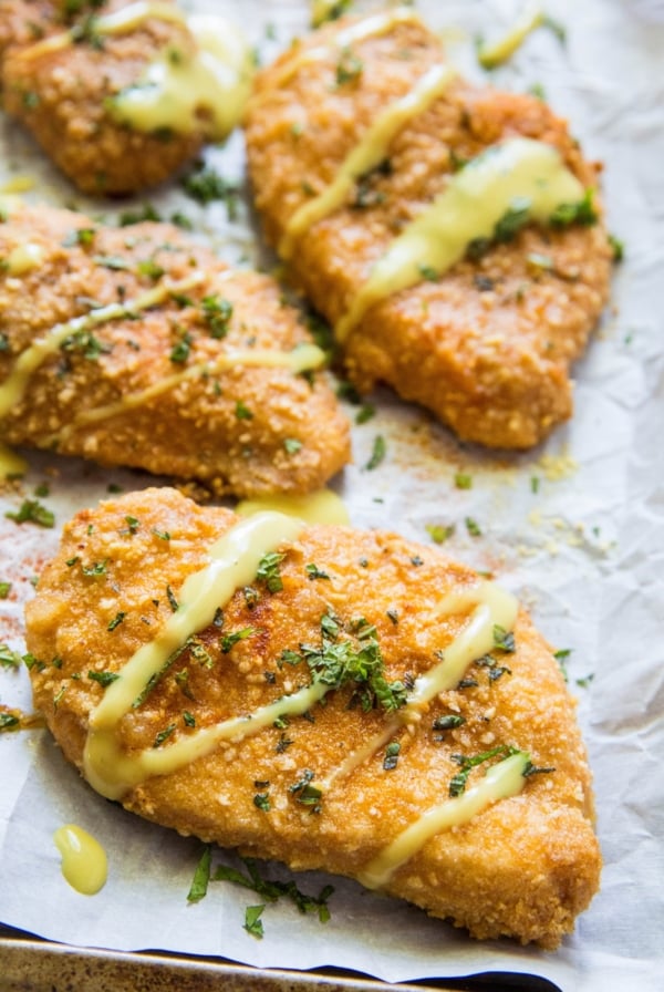 Four filets of Gluten-Free Baked Breaded Chicken on a large baking sheet, drizzled with sauce and sprinkled with fresh parsley.
