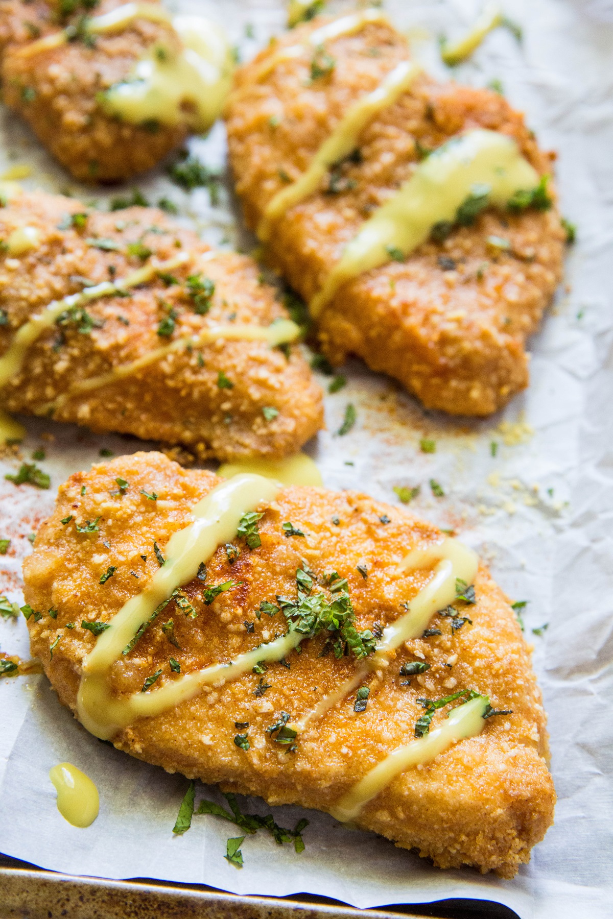 Four filets of Gluten-Free Baked Breaded Chicken on a large baking sheet, drizzled with sauce and sprinkled with fresh parsley.
