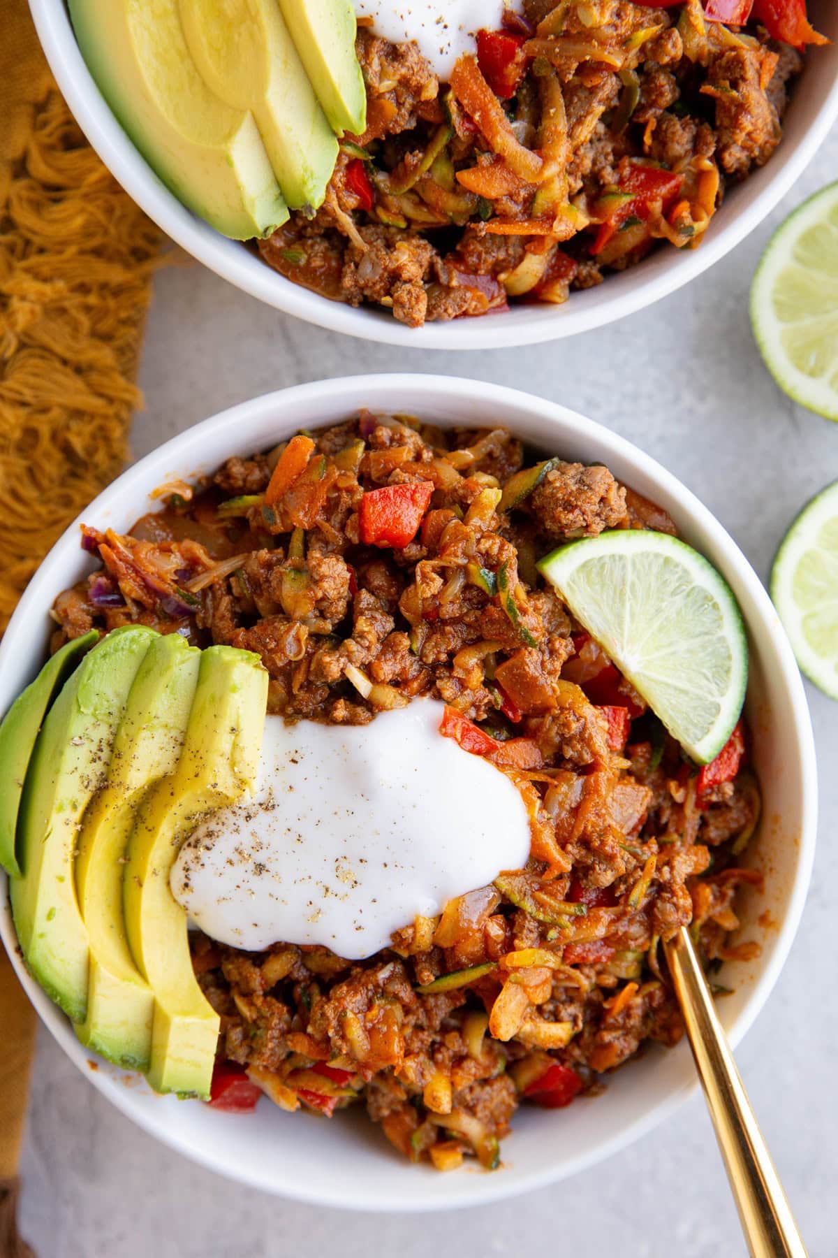 Two white bowls of Mexican ground beef and vegetables with sour cream and avocado on top.