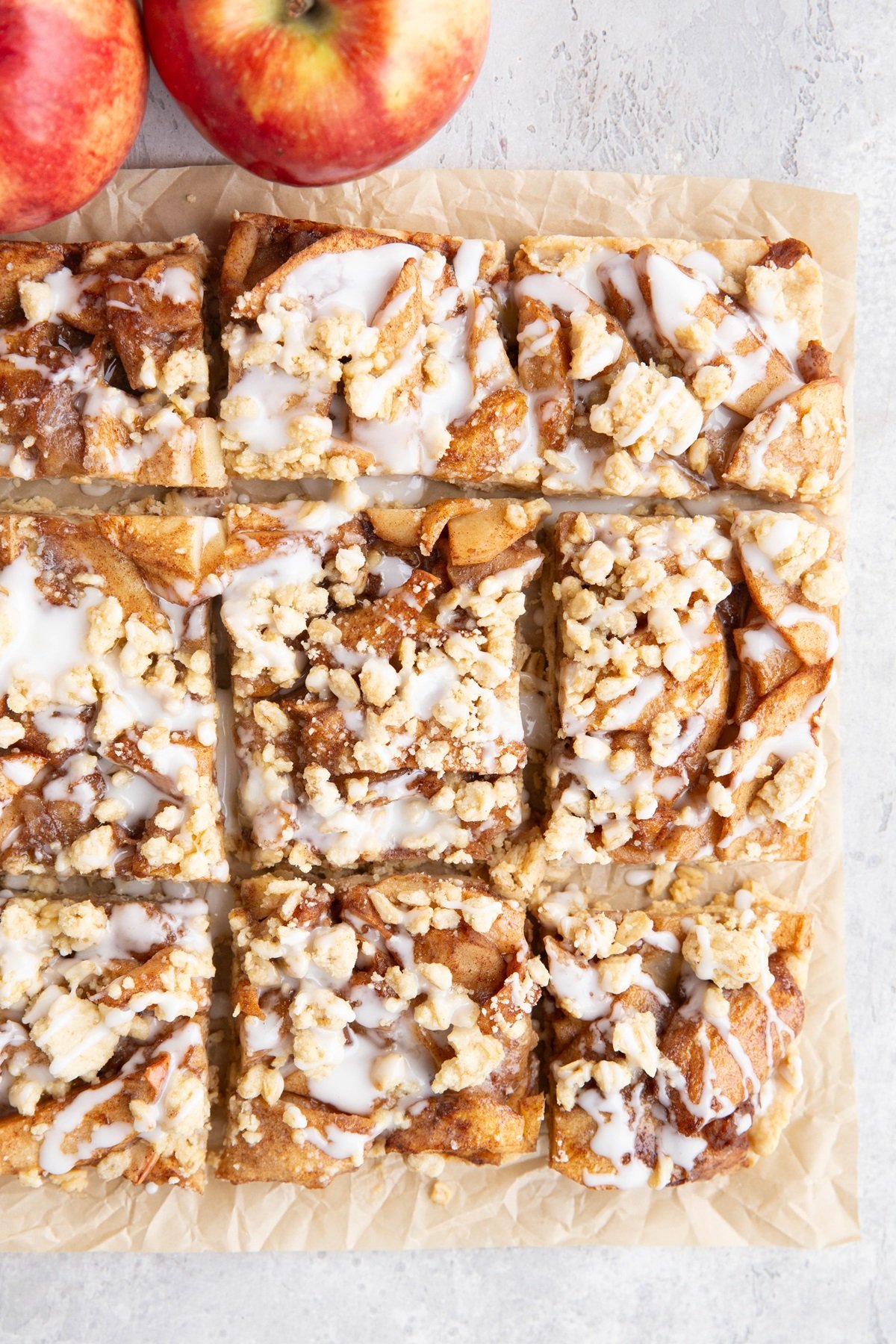Apple crumb bars on a sheet of parchment paper, cut into slices and drizzled with glaze. Fresh apples to the side.