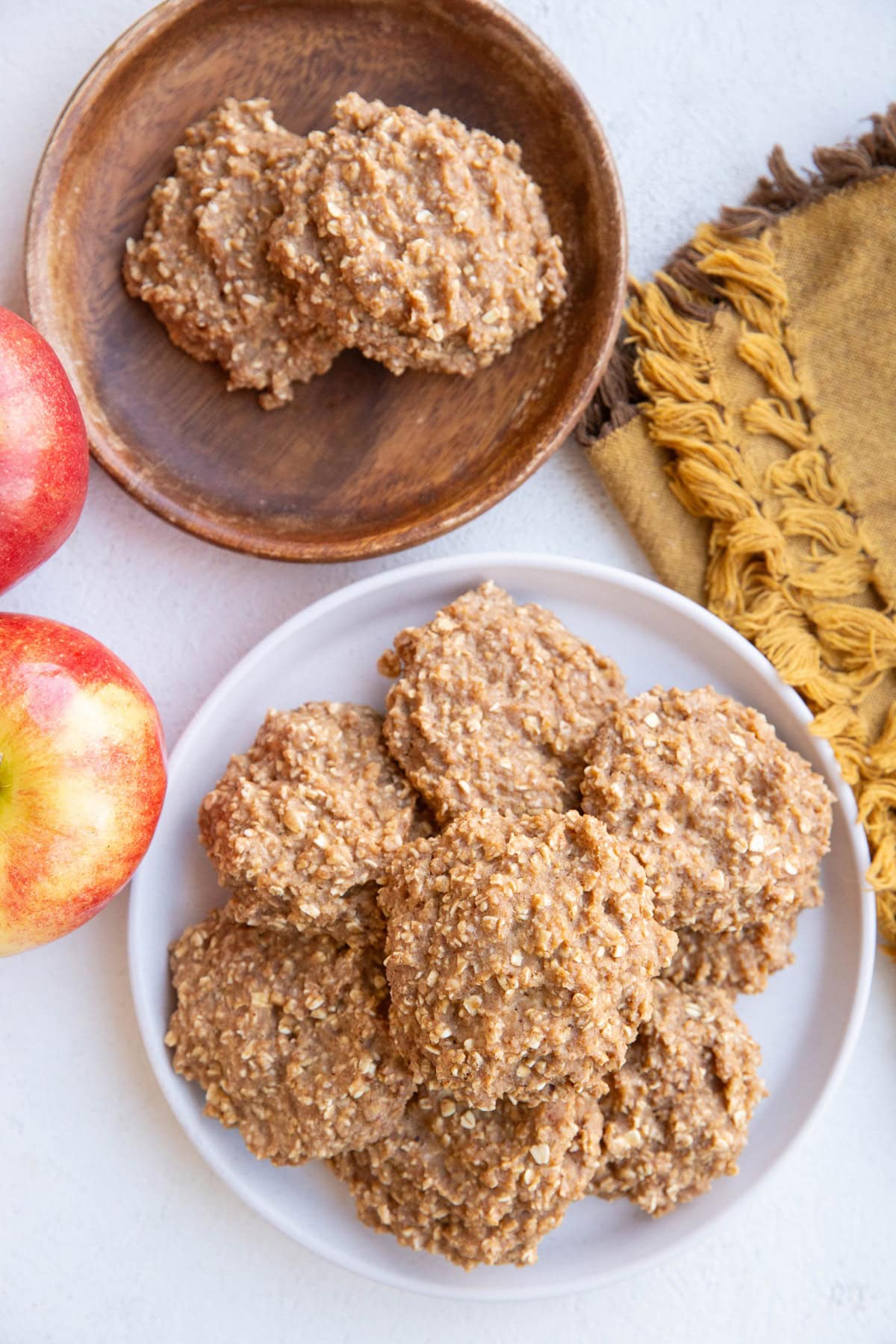 One white plate and one wooden plate with applesauce cookies and a golden napkin and fresh apples to the side.