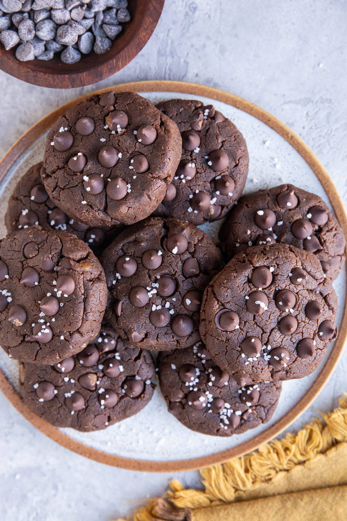 Plate of peanut butter chocolate chickpea cookies sprinkled with sea salt with a napkin to the side and a bowl of chocolate chips.