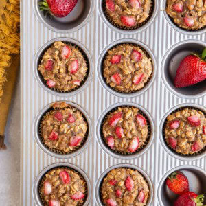 Muffin tray filled with strawberry banana baked oatmeal cups with fresh strawberries and a golden napkin.