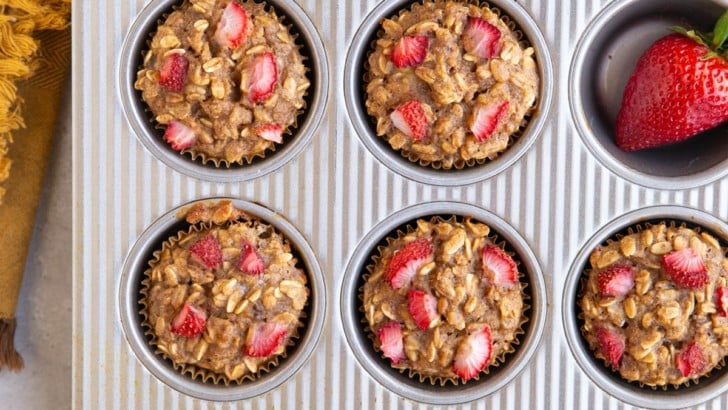 Muffin tray filled with strawberry banana baked oatmeal cups with fresh strawberries and a golden napkin.