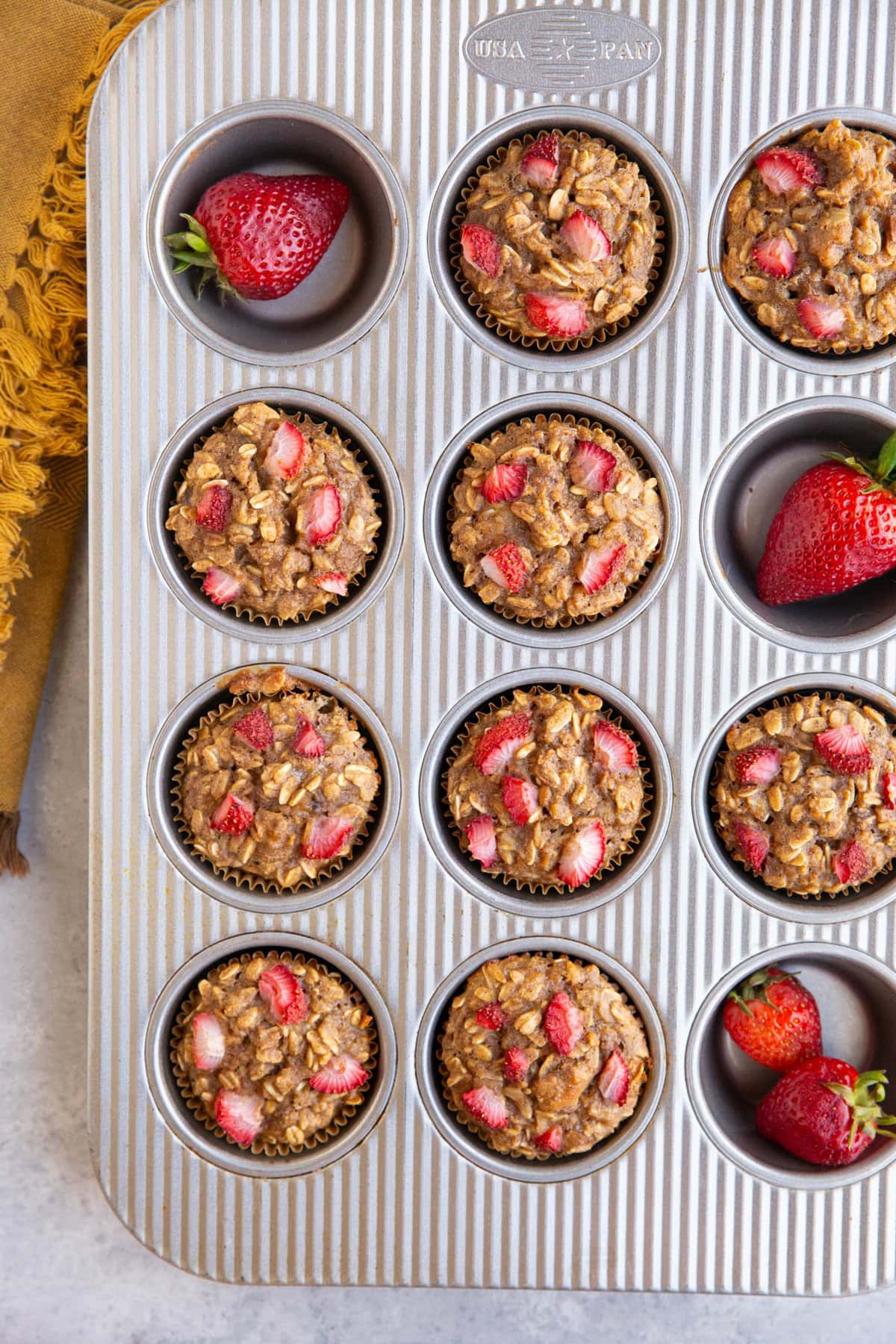 Muffin tray filled with strawberry banana baked oatmeal cups with fresh strawberries and a golden napkin.