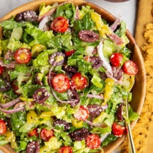 Large bowl of Italian salad with a golden serving spoon, golden napkin, and bowl of cherry tomatoes to the side.