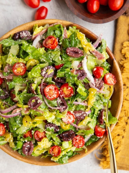 Large bowl of Italian salad with a golden serving spoon, golden napkin, and bowl of cherry tomatoes to the side.