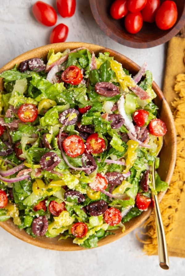 Large bowl of Italian salad with a golden serving spoon, golden napkin, and bowl of cherry tomatoes to the side.