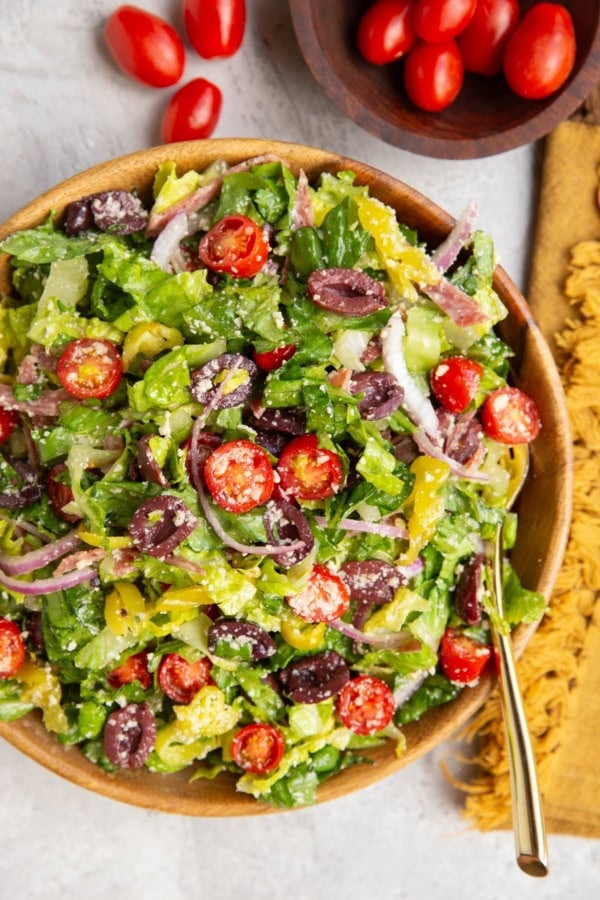 Large bowl of Italian salad with a golden serving spoon, golden napkin, and bowl of cherry tomatoes to the side.