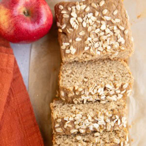 Loaf of applesauce bread on a sheet of parchment paper, cut into slices with fresh apples to the side.
