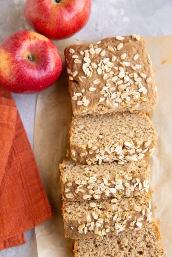 Loaf of applesauce bread on a sheet of parchment paper, cut into slices with fresh apples to the side.