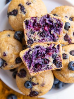 White plate with blueberry muffins and one sliced in half, exposing the blueberry inside.