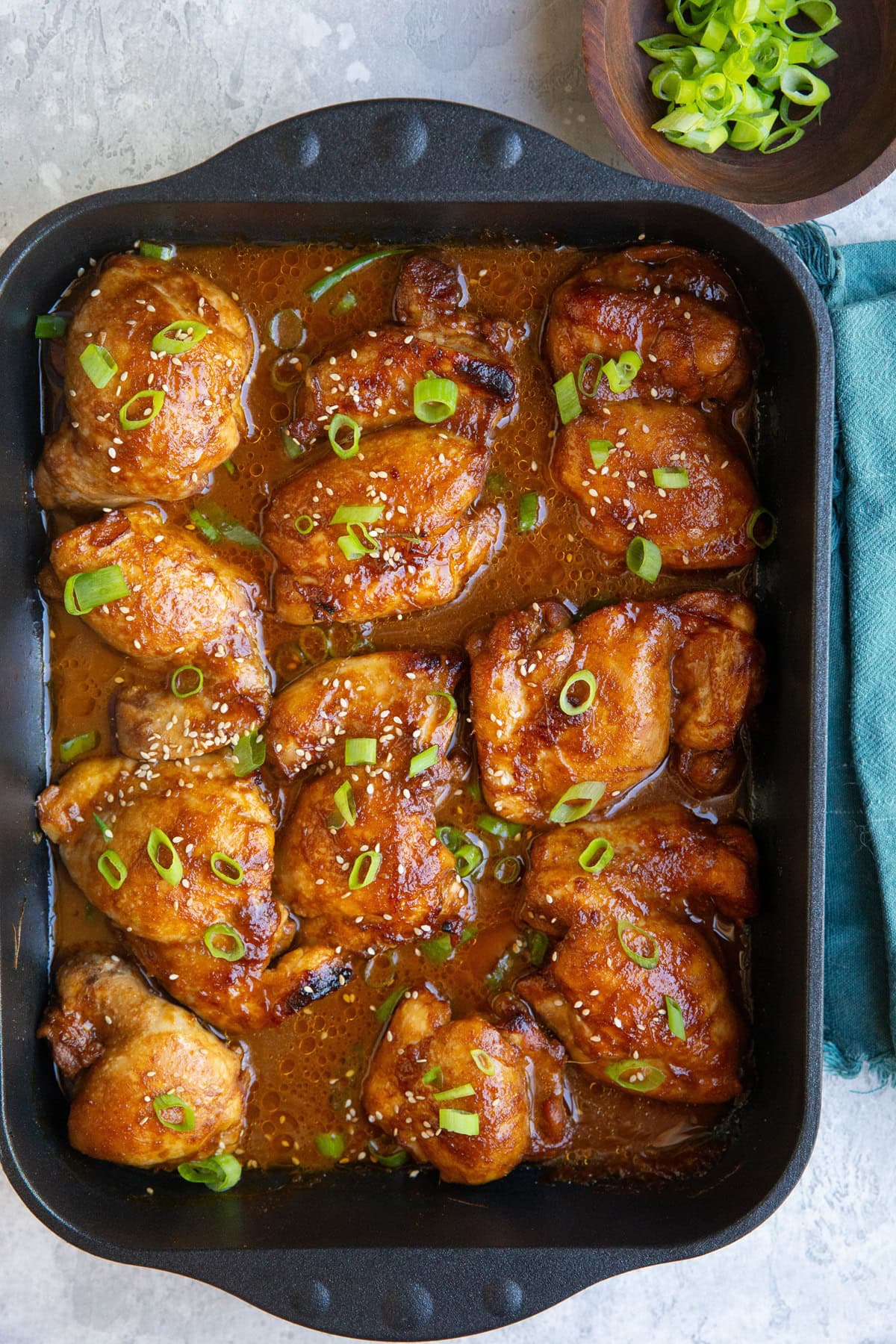 Baking dish full of huli-huli chicken sprinkled with green onions with a bowl of green onions and a blue napkin to the side.