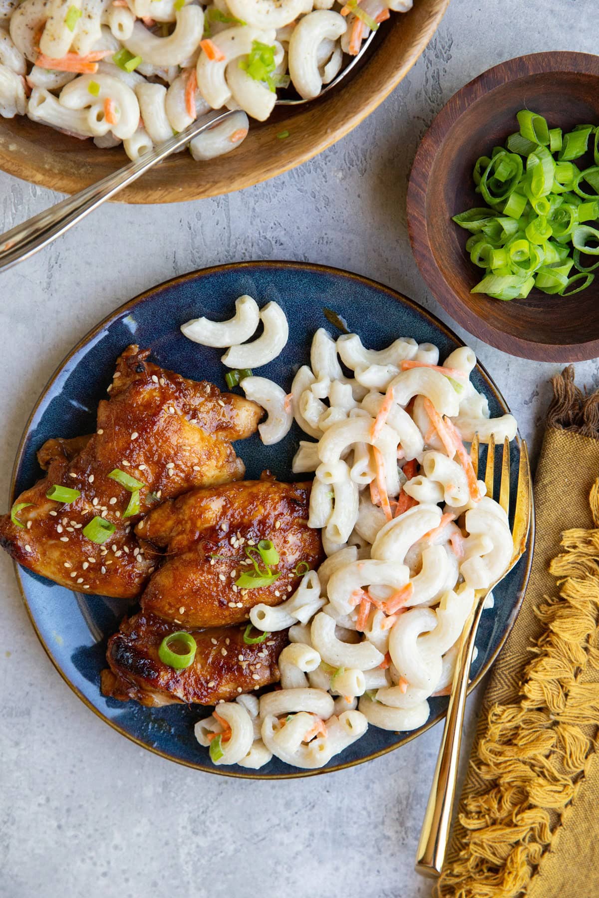 Blue plate of chicken and macaroni salad with a napkin and fork to the side and a bowl of macaroni salad.