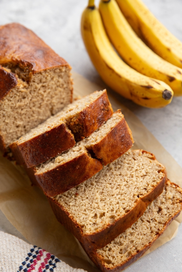 Loaf of high-protein banana bread on a backdrop, cut into slices with bananas in the background.