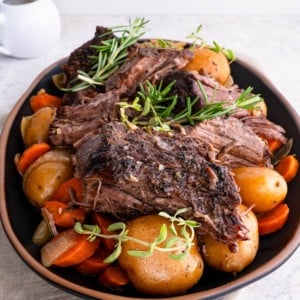 Platter with pot roast on top of potatoes and carrots, ready to serve with a vessel of gravy in the background.