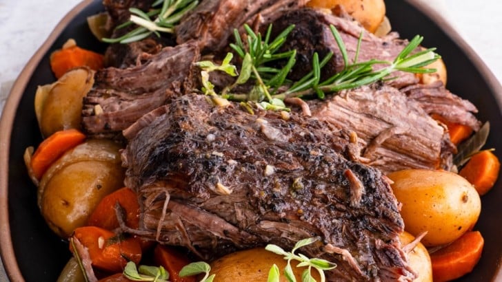 Platter with pot roast on top of potatoes and carrots, ready to serve with a vessel of gravy in the background.