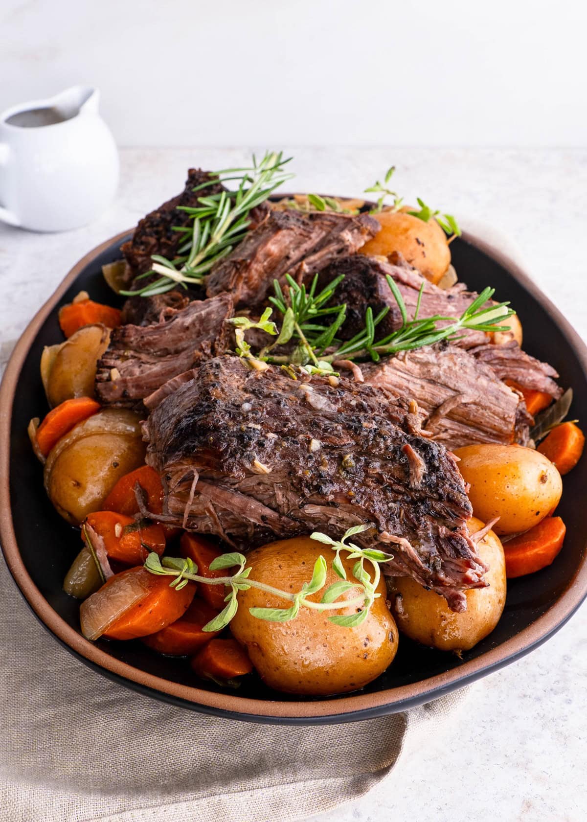 Platter with pot roast on top of potatoes and carrots, ready to serve with a vessel of gravy in the background.