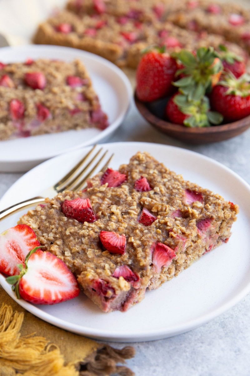 Two slices of strawberry breakfast bars on white plates with the rest of the breakfast bars in the background.