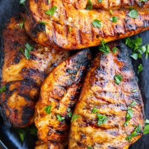 Black plate with four grilled chicken breasts sprinkled with parsley and slices of lemon to the side.