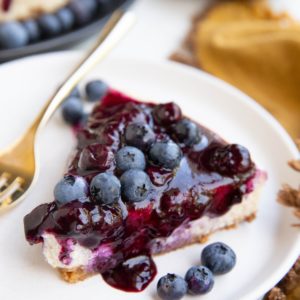 Slice of blueberry cheesecake on a white plate with the rest of the cheesecake in the background.
