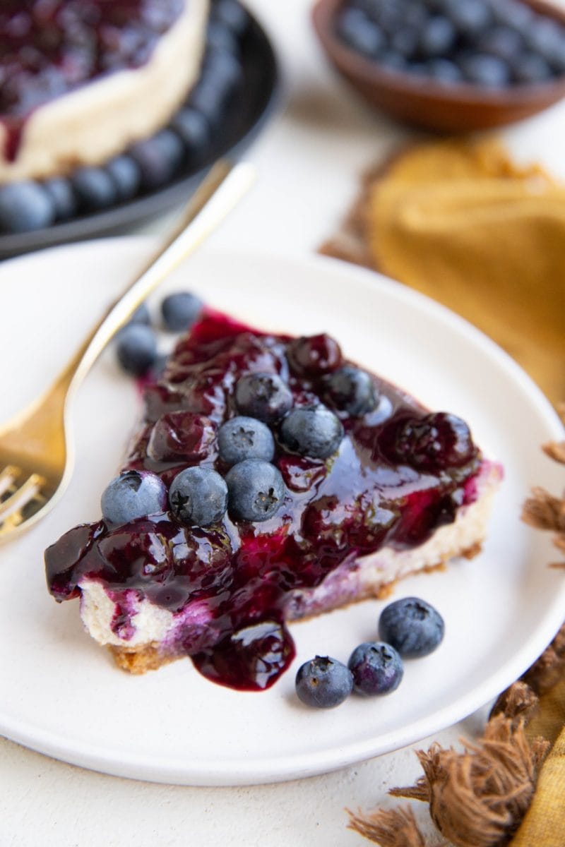 Slice of blueberry cheesecake on a white plate with the rest of the cheesecake in the background.