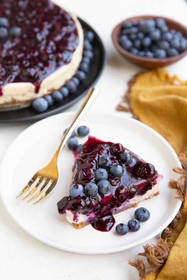 Slice of blueberry cheesecake on a plate with the rest of the blueberry cheesecake in the background.