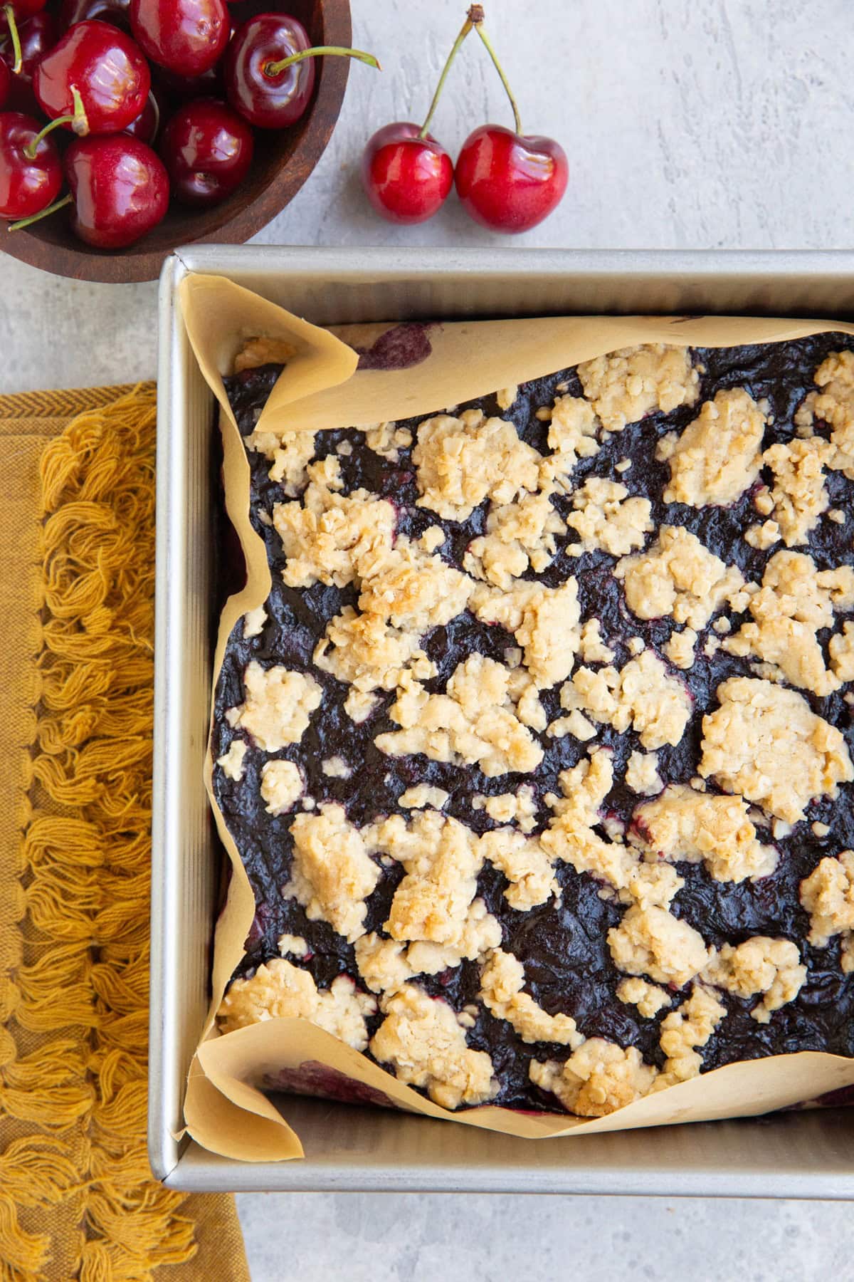 Baking pan with cherry pie bars inside fresh out of the oven and a bowl of fresh cherries to the side.
