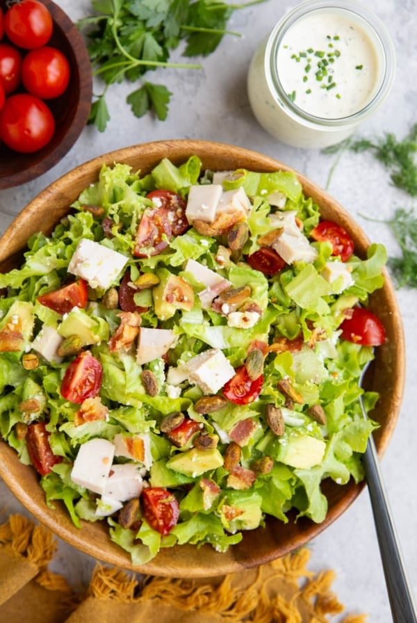 Chicken bacon avocado ranch salad in a wooden bowl with ranch dressing to the side and a bowl of tomatoes to the side.