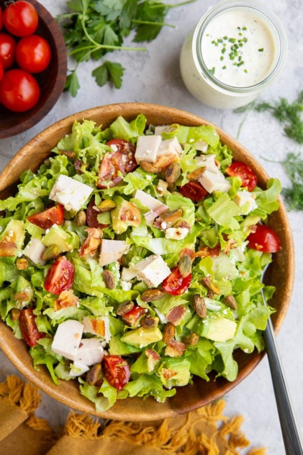 Chicken bacon avocado ranch salad in a wooden bowl with ranch dressing to the side and a bowl of tomatoes to the side.