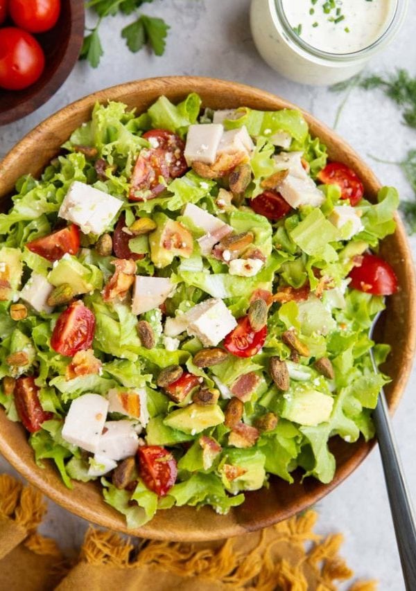 Chicken bacon avocado ranch salad in a wooden bowl with ranch dressing to the side and a bowl of tomatoes to the side.