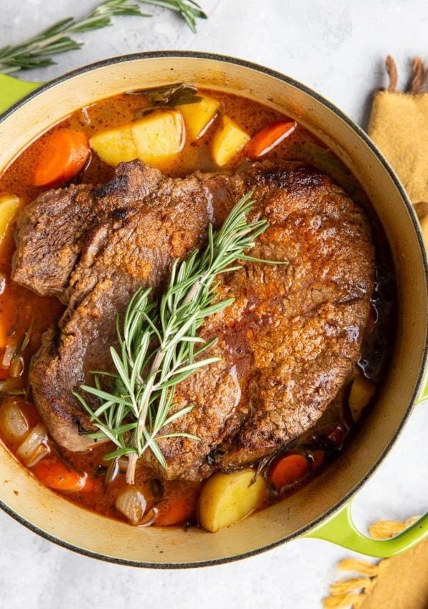 Pot roast in a Dutch oven with fresh herbs and vegetables.