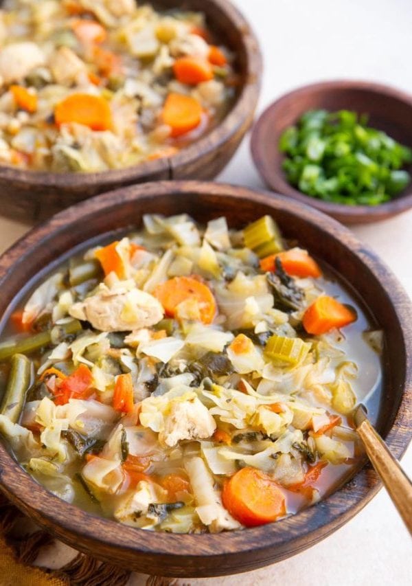 Chicken and cabbage soup in two wooden bowls with a bowl of green onions to the side, ready to eat.