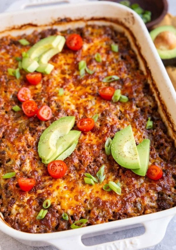Casserole dish full of cheesy enchilada casserole topped with avocado and tomatoes