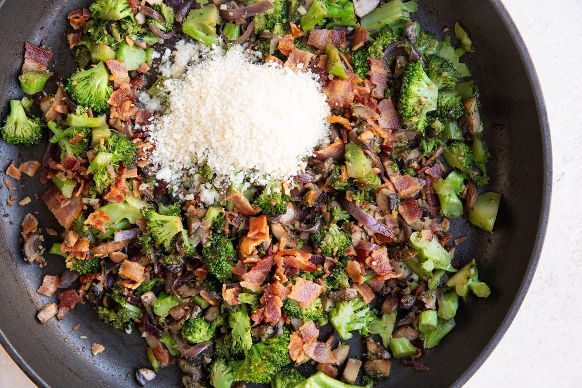 Parmesan cheese and crispy bacon on top of broccoli in a skillet to be mixed in.
