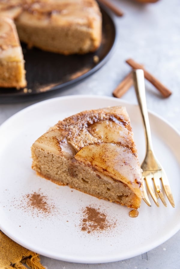 Slice of pear cake on a white plate with gold fork and cake in the background.