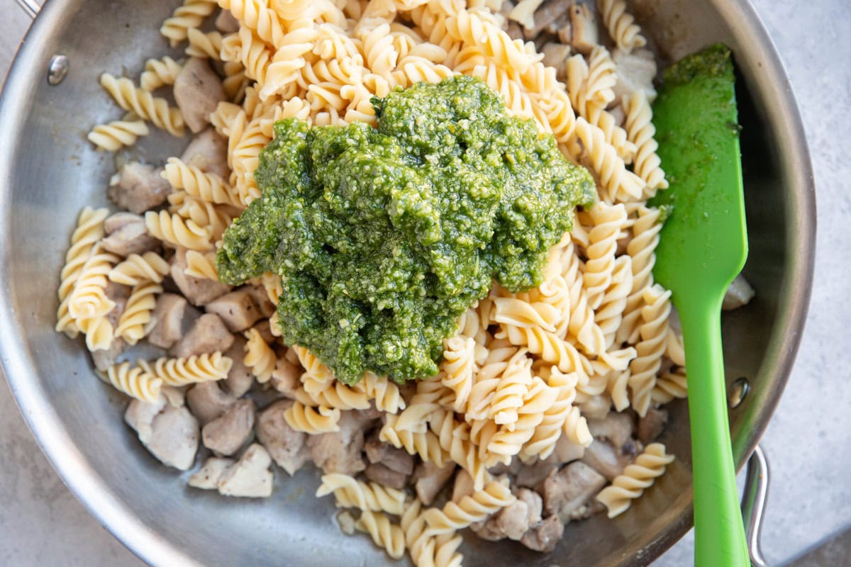 Cooked chicken, pasta, and pesto sauce in a skillet, ready to be stirred together.