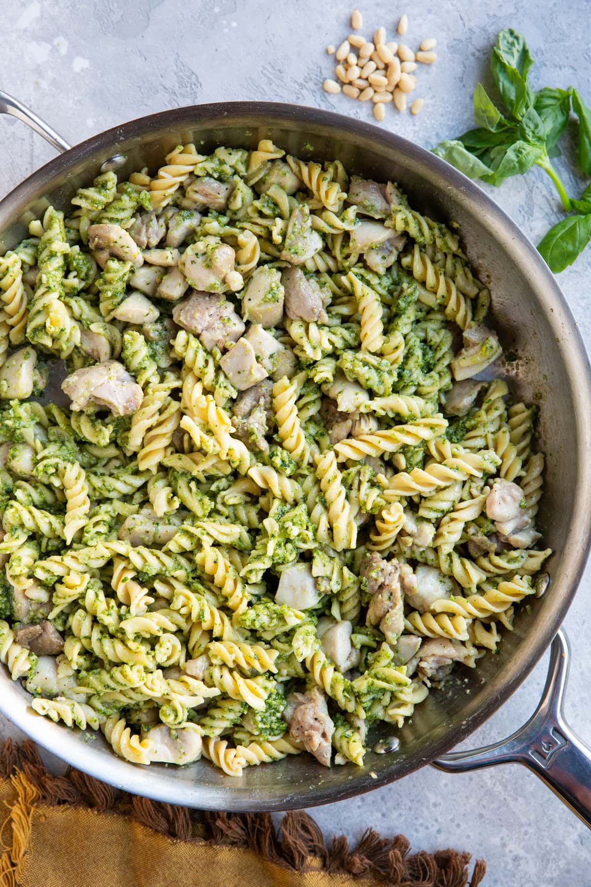 Skillet of chicken pesto pasta, ready to serve. Fresh basil and pine nuts to the side of the skillet.